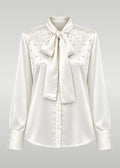 An ivory satin blouse with a large bow tie at the collar, buttoned front, and delicate bead embellishments across the yoke, shown on a neutral gray gradient background.
