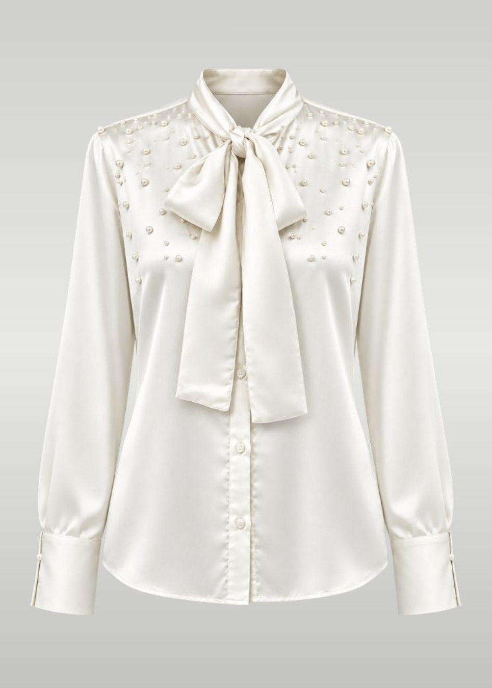 An ivory satin blouse with a large bow tie at the collar, buttoned front, and delicate bead embellishments across the yoke, shown on a neutral gray gradient background.