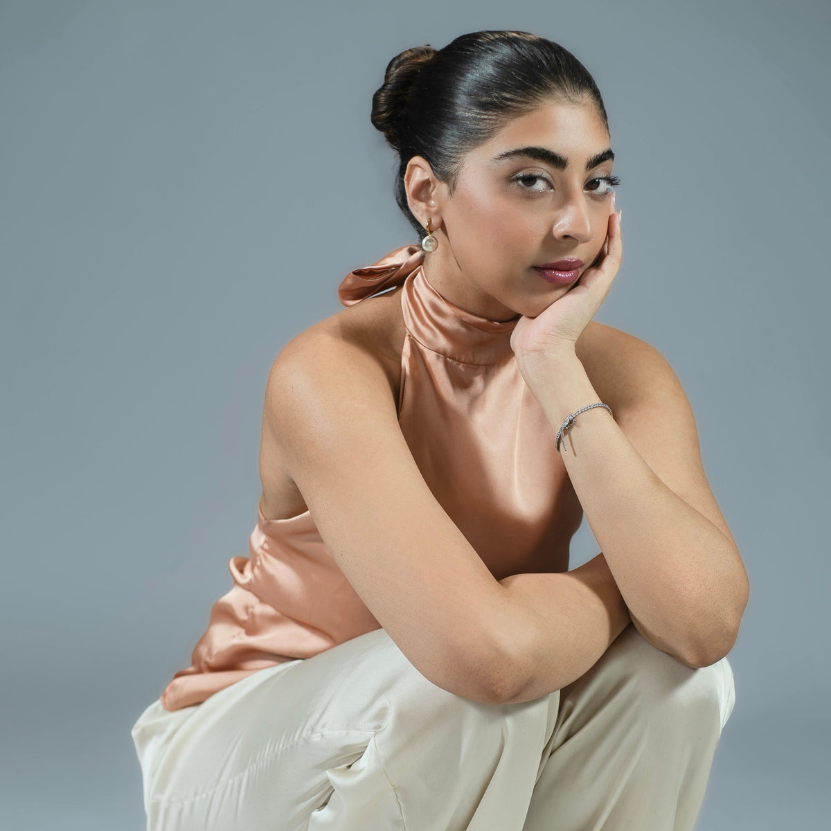 A woman crouching in a studio setting, wearing a sleeveless peach satin top and cream wide-leg pants against a solid gray background.